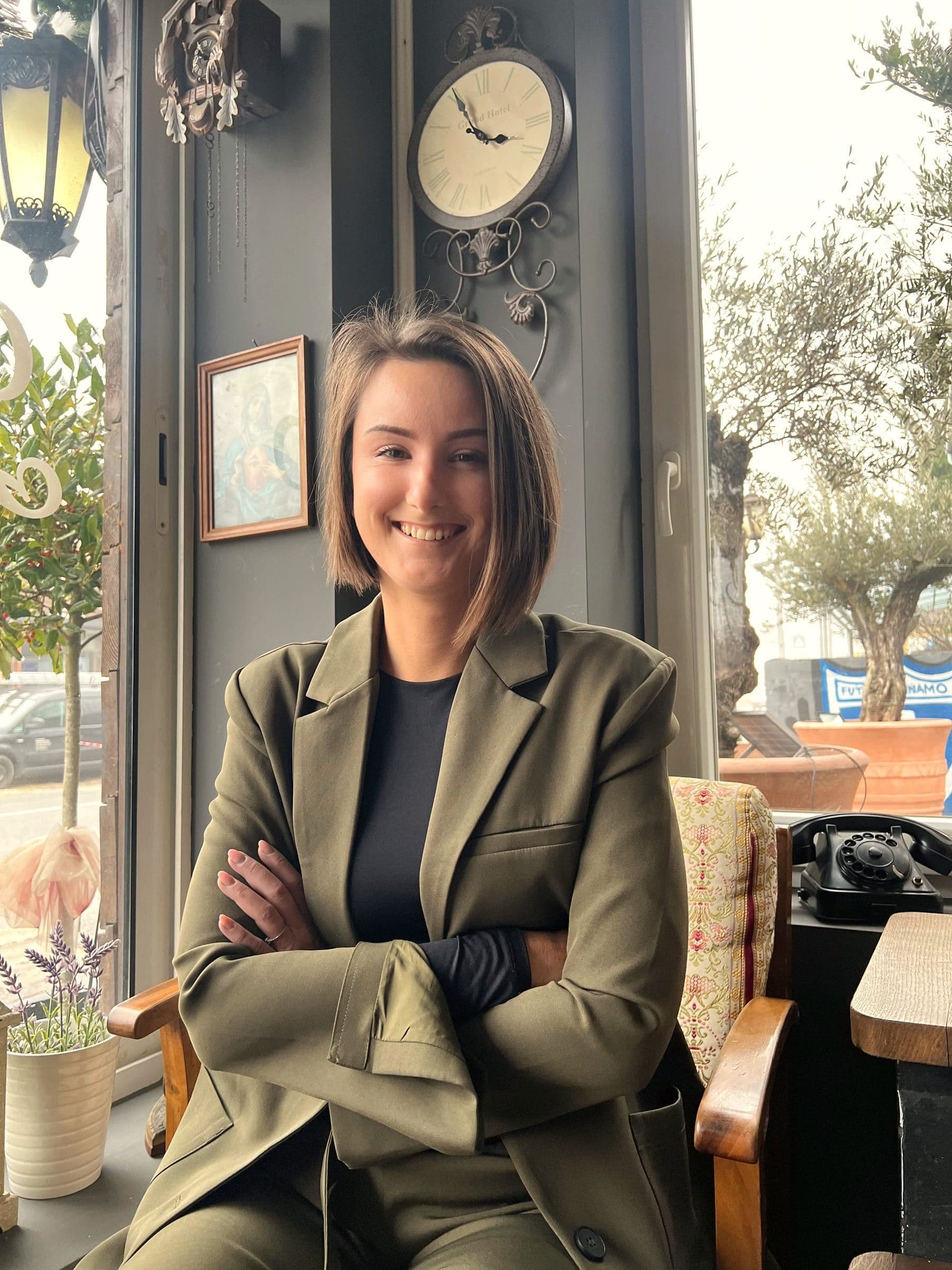 Adrijana u maslinastom odijelu sjedi prekriženih ruku i smiješi se u kameru, iza nje prozor kafića s maslinama.