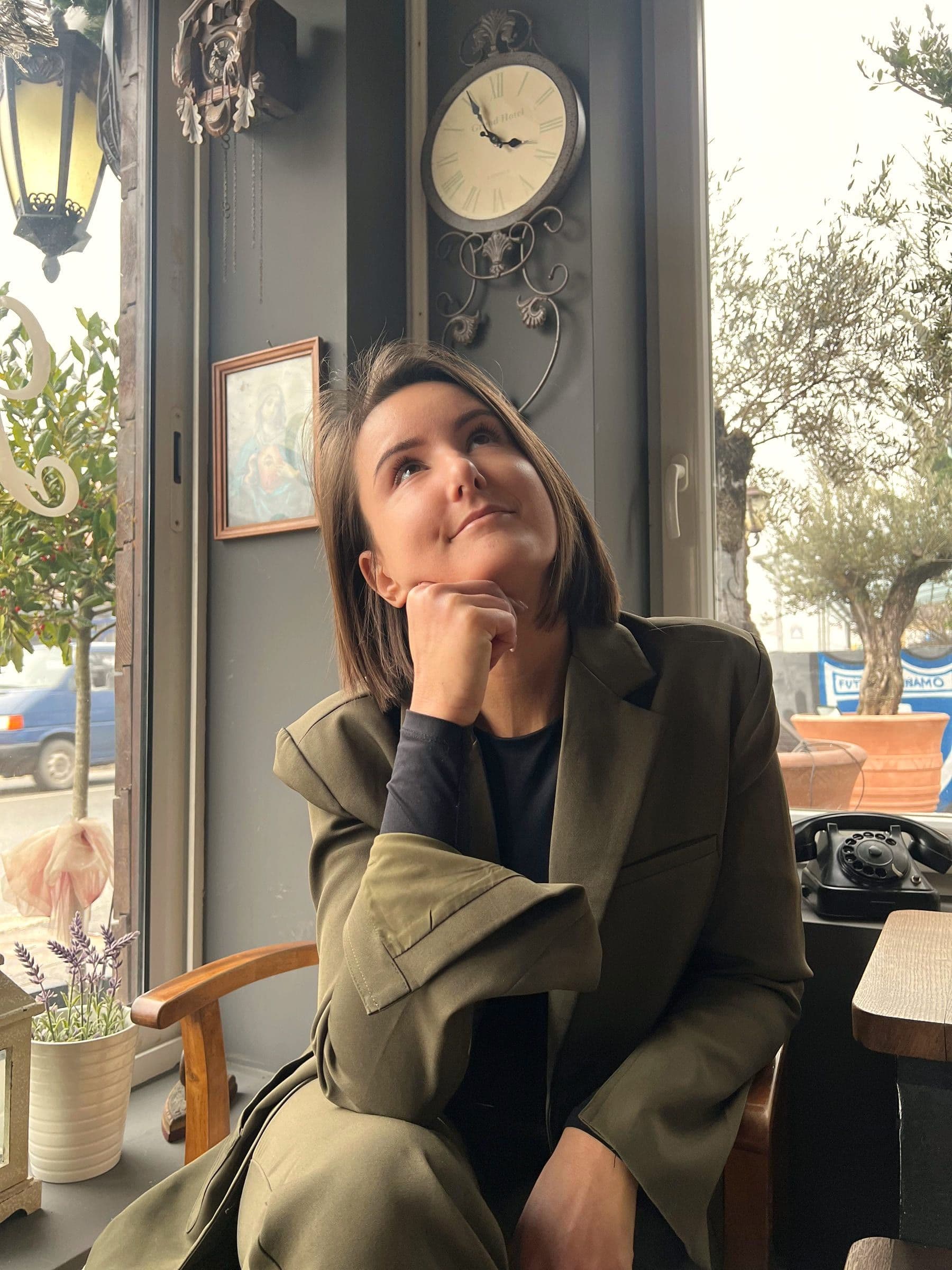 Adrijana naslonjene glave na ruku gleda prema gore, zamišljena, u kafiću sa zidnim satom iza sebe.